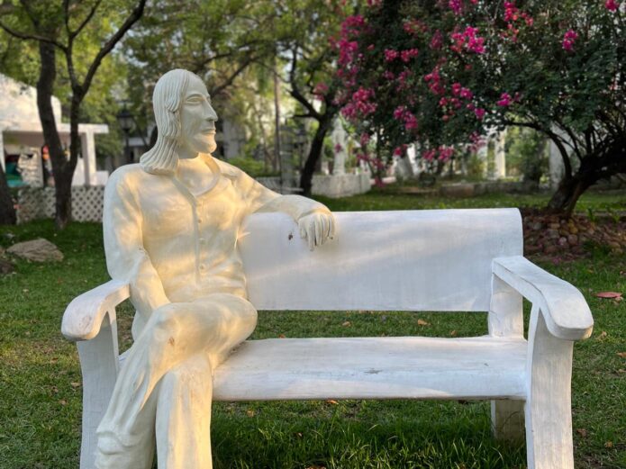 White sculpture of a seated figure on a garden bench, gazing sideways, with bougainvillea and trees in the background.