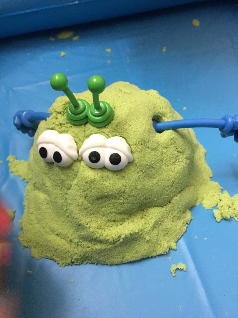 Green kinetic sand creature with googly eyes and antennae, looking mildly bewildered