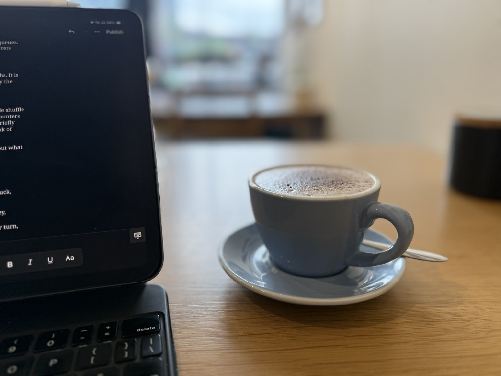 Coffee in a grey ceramic cup beside an iPad showing a draft in progress, on a wooden café table