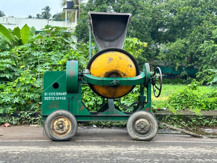 A green and yellow concrete mixer on a wet road in Kerela, surrounded by lush vegetation.
