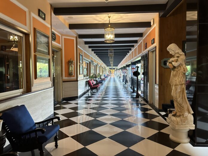 A long hotel corridor in India with a black and white chequered floor, ochre walls lined with framed paintings, brass pendant lights, and a marble statue in the foreground.