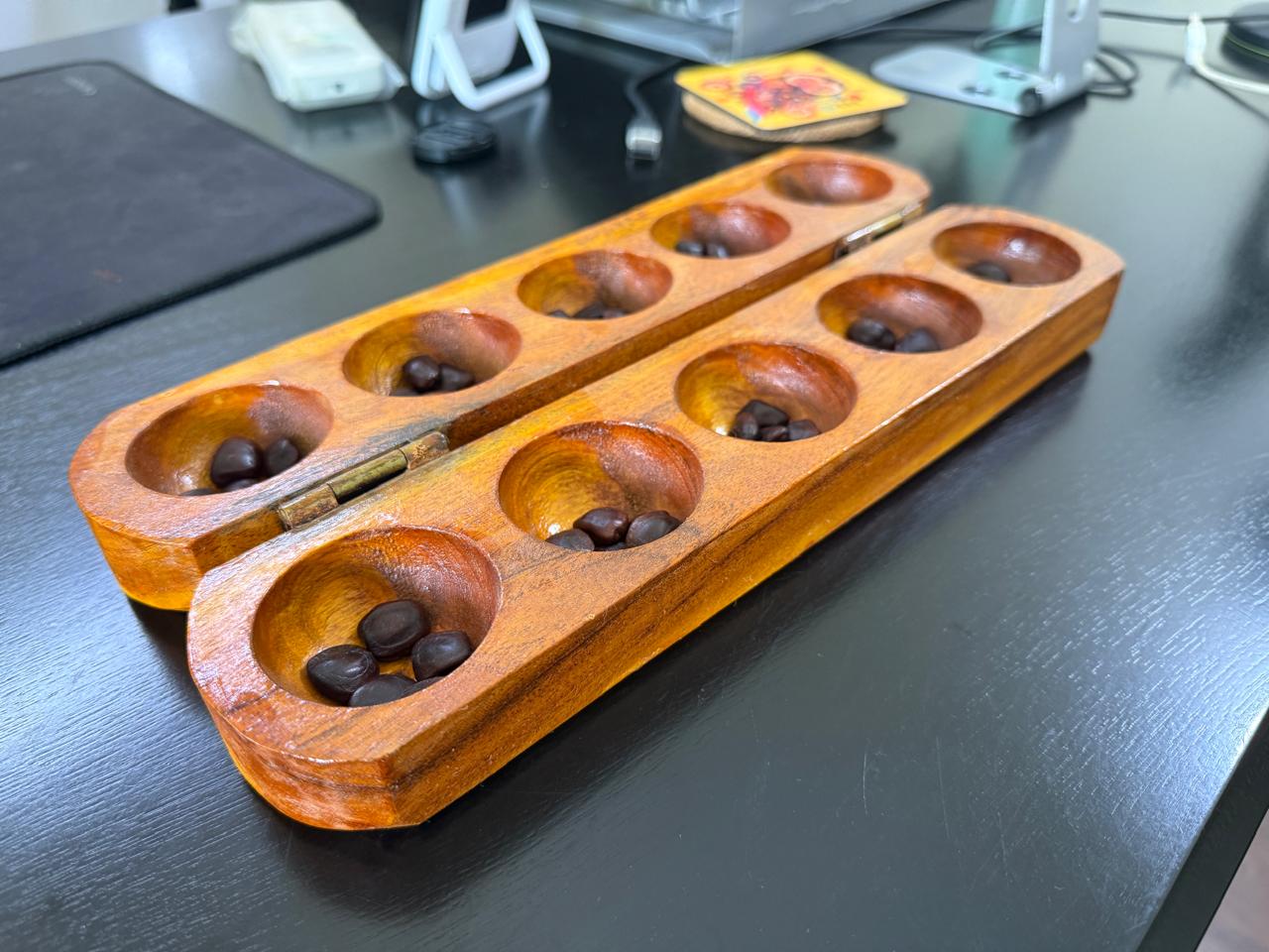 A wooden pallanguli board with tamarind seeds in carved hollows, placed on a dark desk.
