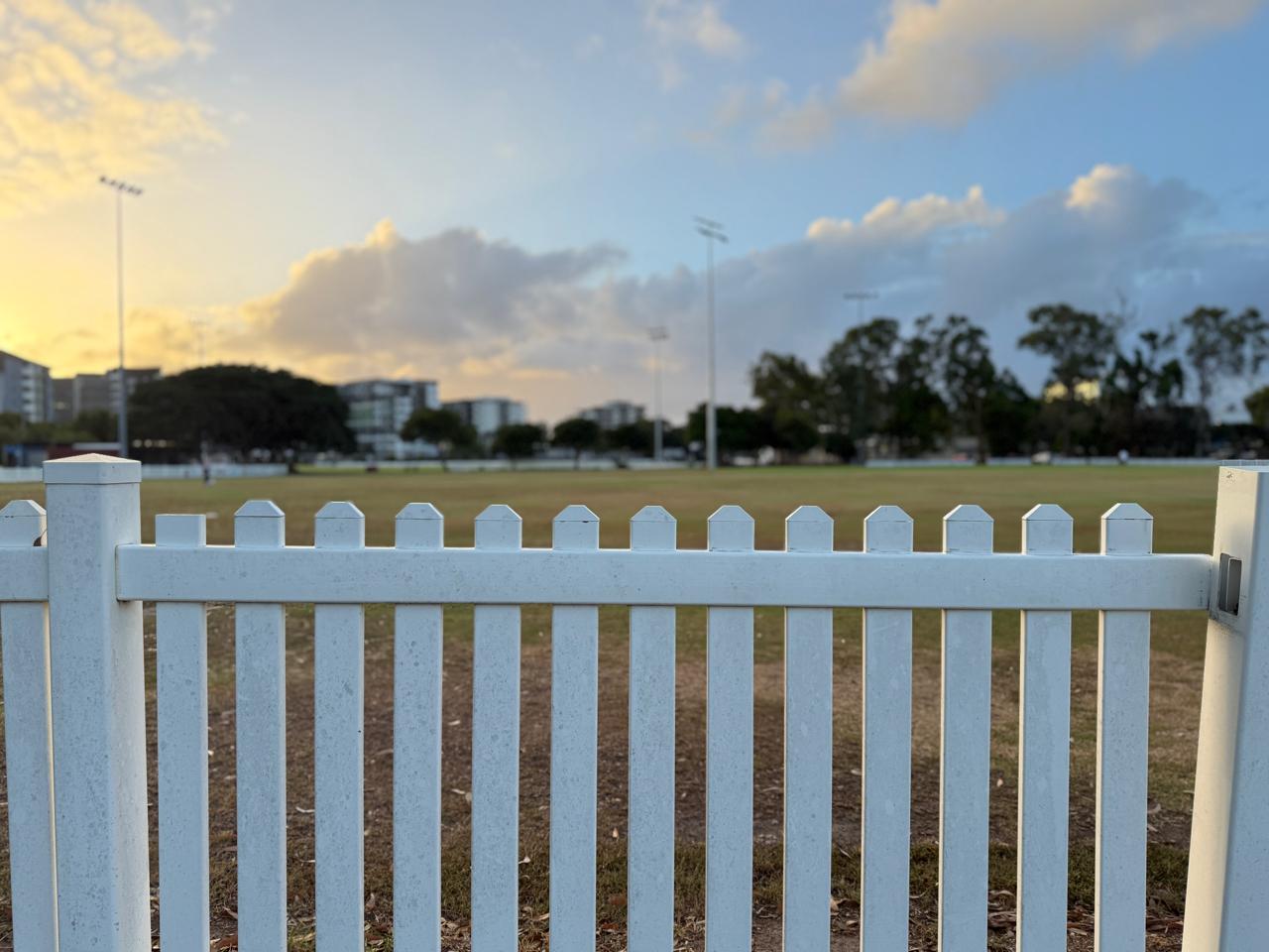 An empty cricket ground at dusk, white picket fence in the foreground, floodlights and city skyline behind