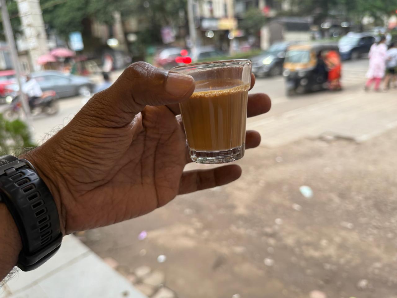 A hand holding a glass of cutting chai on a Mumbai street, with traffic and an auto rickshaw behind.