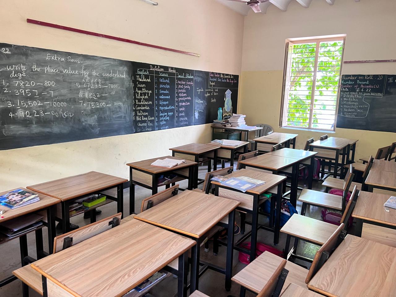 An empty primary school classroom with wooden desks and a long blackboard. The board carries place value sums, a dictation list of Southeast Asian place names, science and social studies words, and an attendance count of twenty pupils.